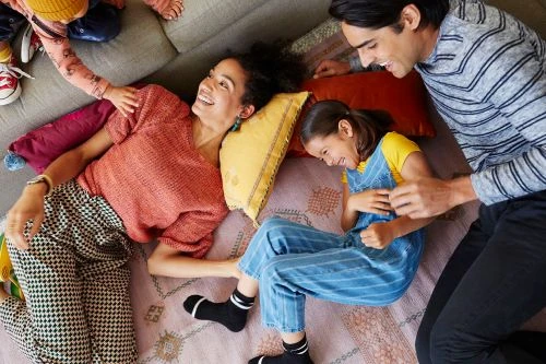 Family of four relaxes together on a patterned rug in a home living room, with two adults and two children, all smiling and playful.