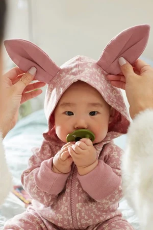 Baby wearing a bunny costume with floral print and a green pacifier, seated indoors and hands adjusting the ears of the bunny hood.