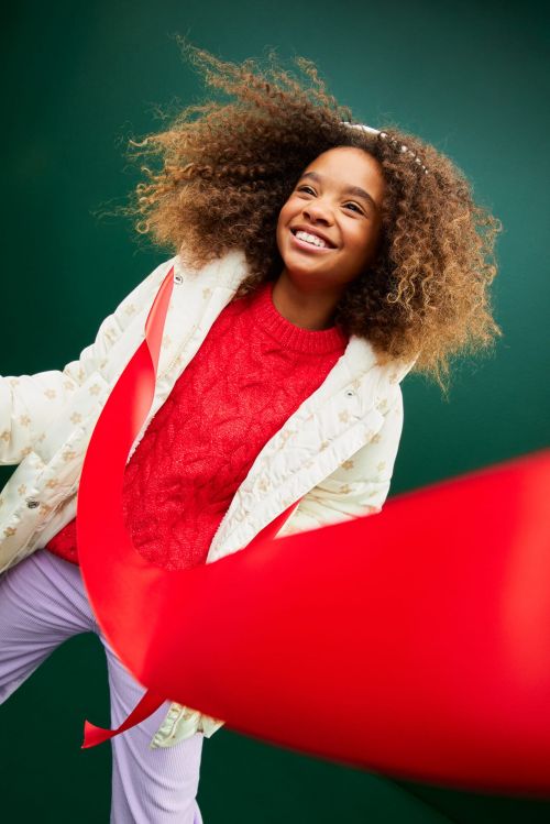 Smiling child with afro hair wearing a cream floral jacket, red sweater, and lavender pants holding a large red ribbon against a green background.