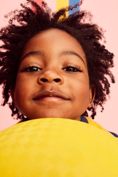 Close-up portrait of an African-American child with curly hair, looking directly at the camera against a pink backdrop with color stripes.
