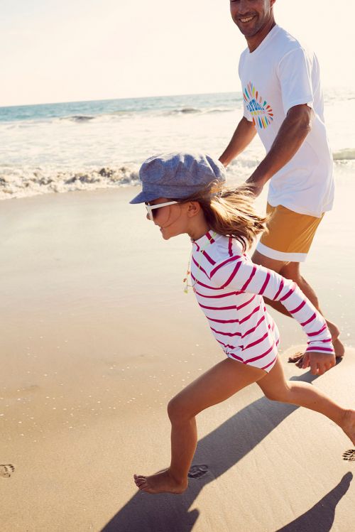A young girl in a striped rash guard runs barefoot on a sandy beach with a smiling adult male in the background, near ocean waves.