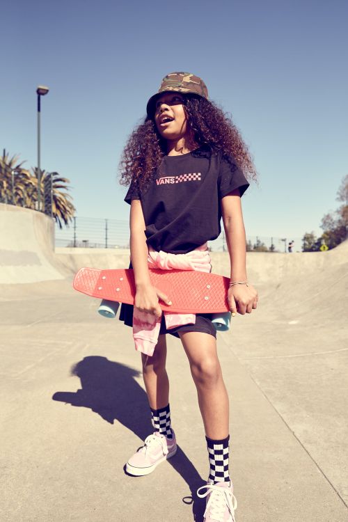 Girl in bucket hat carries skateboard at skatepark in black graphic tee, patterned socks, and trainers for sport and fashion.