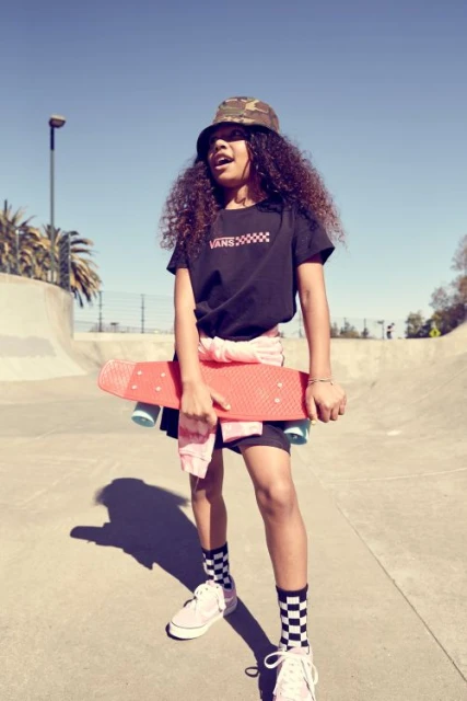 Girl in bucket hat carries skateboard at skatepark in black graphic tee, patterned socks, and trainers for sport and fashion.