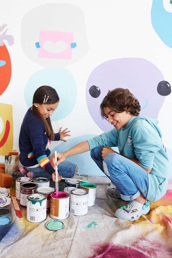 Two children are painting a mural on a wall indoors. They are surrounded by paint cans, paintbrushes, and a paint-splattered drop cloth.