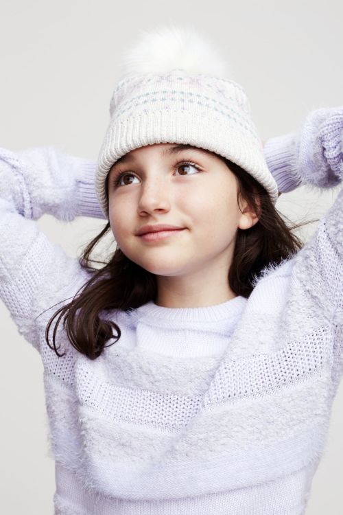 Child wearing a white pom-pom beanie and lavender knitted sweater against a plain background