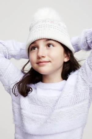 Child wearing a white pom-pom beanie and lavender knitted sweater against a plain background