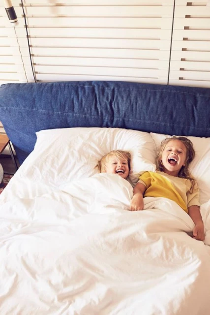 Two happy young children are laughing and lying in a bed with white linens and a denim-style headboard, captured in a bright indoor setting.