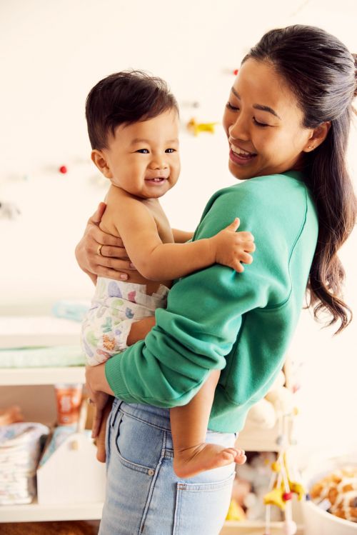 Smiling mother wearing a green sweatshirt and jeans holding her baby who is wearing a diaper in an indoor setting.