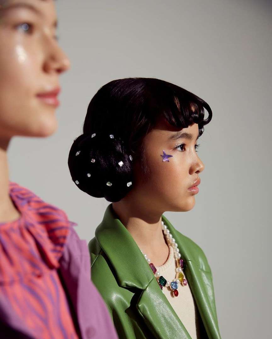 Two young models pose in a studio, showcasing fashion; one wears a green jacket and pearl necklace, the other, a colorful top.