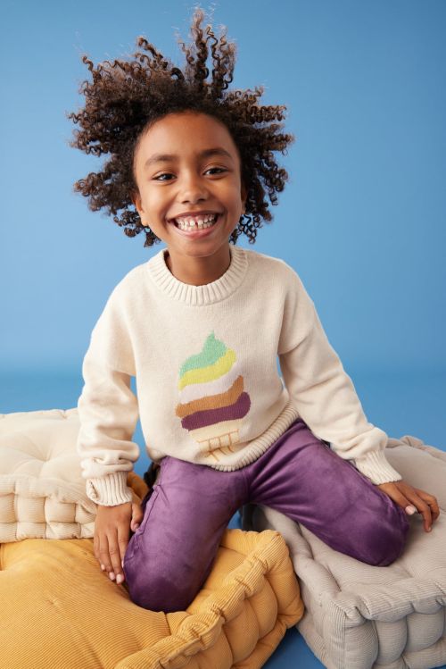 Smiling child with curly hair poses in a cream-colored sweater with a graphic, purple pants, and surrounded by floor cushions against blue.