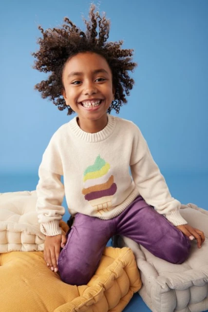 Smiling child with curly hair poses in a cream-colored sweater with a graphic, purple pants, and surrounded by floor cushions against blue.