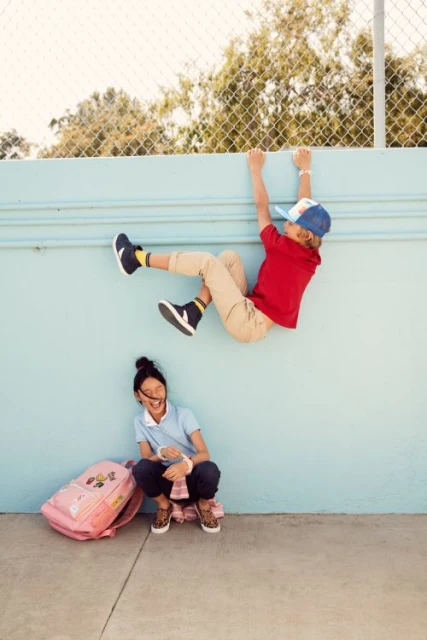 Two children at play. One boy hangs from a wall, while the other child sits below with a backpack, smiling on a sunny day.