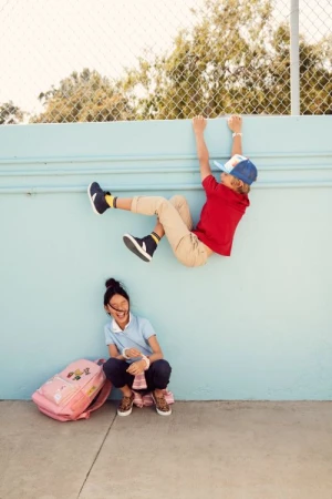 Two children at play. One boy hangs from a wall, while the other child sits below with a backpack, smiling on a sunny day.
