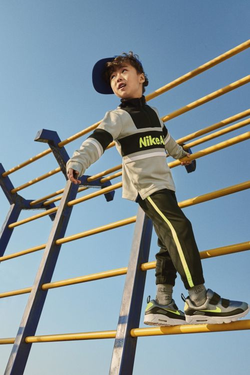 A young boy wearing a Nike outfit and a blue baseball cap climbs a yellow and blue jungle gym against a clear blue sky.