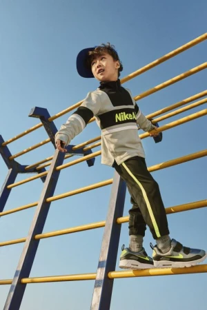 A young boy wearing a Nike outfit and a blue baseball cap climbs a yellow and blue jungle gym against a clear blue sky.