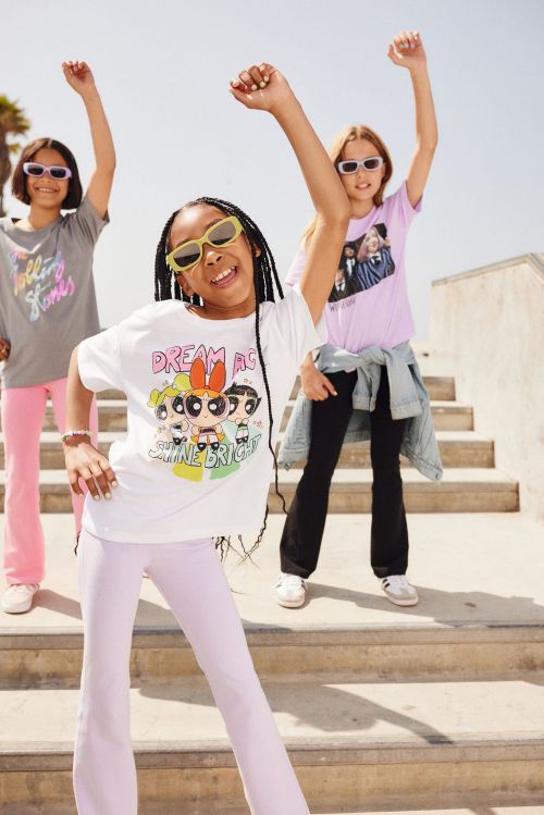 Three young girls pose in sunglasses and graphic tees on concrete stairs, smiling with arms raised in an outdoor, daytime setting.