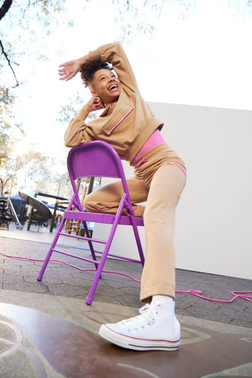 A woman in a tan outfit poses on a purple folding chair outdoors, smiling with one arm overhead and white high-top sneakers.