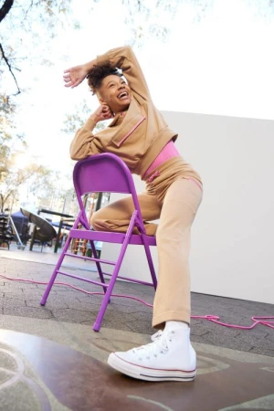A woman in a tan outfit poses on a purple folding chair outdoors, smiling with one arm overhead and white high-top sneakers.