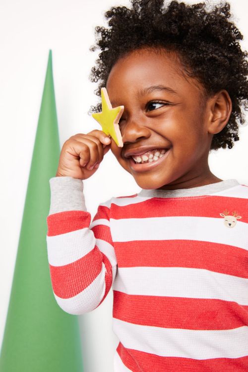 Young child in striped shirt holds a yellow star up to their eye indoors with a minimalist Christmas tree in the background.