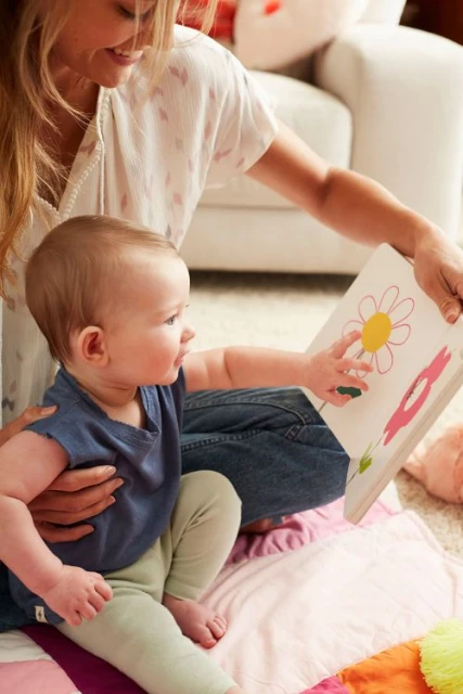 Young child and adult are sitting on a quilt while looking at a picture book with flower illustrations in an indoor living room setting.