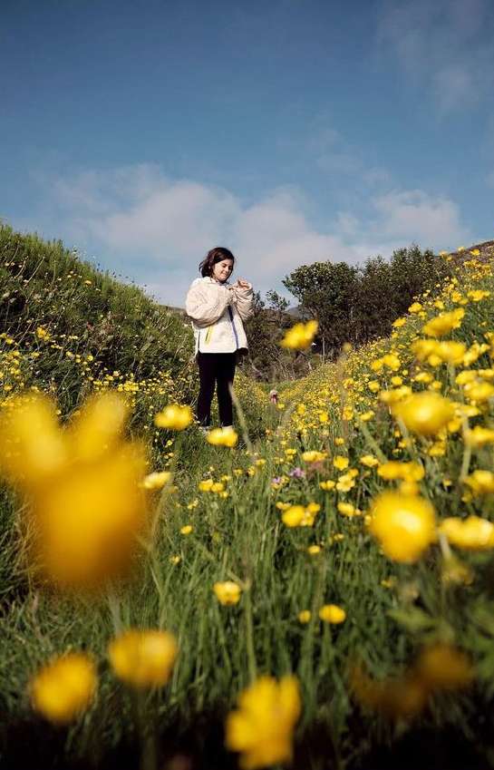 Young woman stands in a field of yellow wildflowers on a hillside under a blue sky with light clouds in a rural outdoor setting.