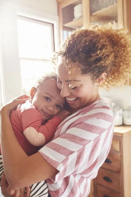 Mother smiling and holding her baby close in a bright, naturally lit kitchen. They are both indoors and wearing striped shirts.