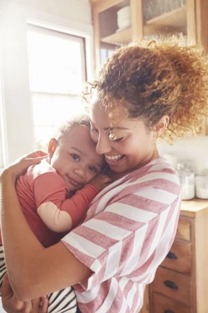 Mother smiling and holding her baby close in a bright, naturally lit kitchen. They are both indoors and wearing striped shirts.
