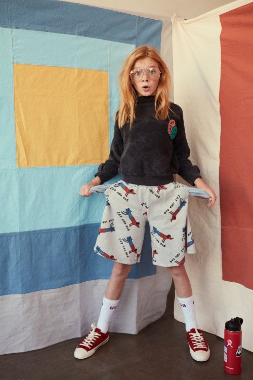 A young child model with red hair poses in a dark sweatshirt and printed shorts, pulling out their pockets in a studio setting.