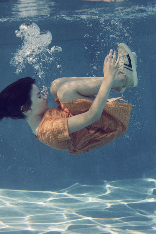 Young woman in an orange dress floating underwater in a pool with bubbles and light reflections creating a tranquil, surreal scene.