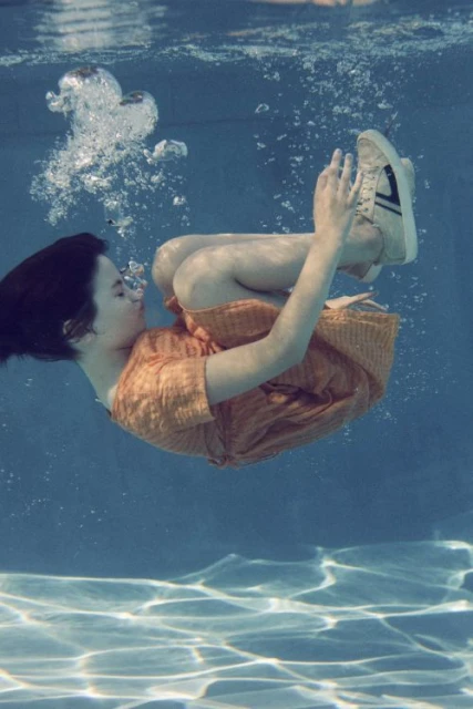 Young woman in an orange dress floating underwater in a pool with bubbles and light reflections creating a tranquil, surreal scene.