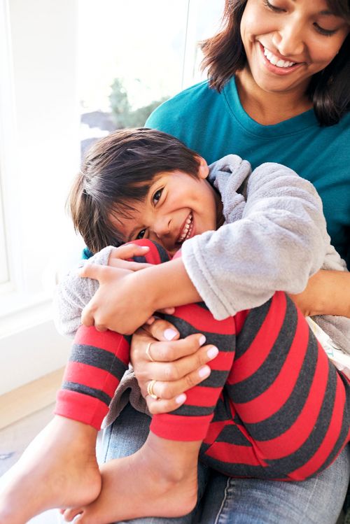 Smiling child with striped leggings is held on the lap of a smiling adult female near a window with natural light and indoor setting.