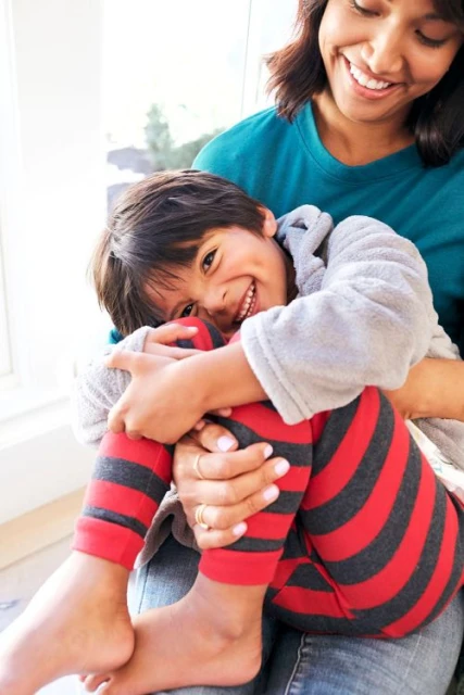 Smiling child with striped leggings is held on the lap of a smiling adult female near a window with natural light and indoor setting.