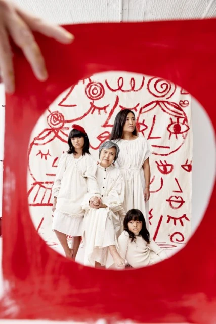 Portrait of a four generation Asian family in white outfits posing in front of a graphic art backdrop framed by red art board.
