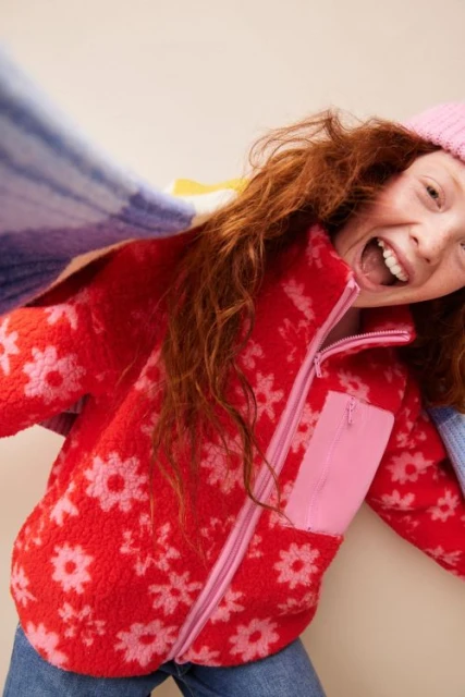 Cheerful young girl wearing a red floral fleece jacket and pink beanie smiles with her mouth open in an indoor setting.