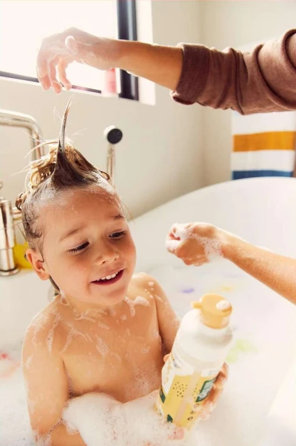 A young child with wet hair stands in a bathtub full of bubbles, with an adult adding soap to the child's wet hair.