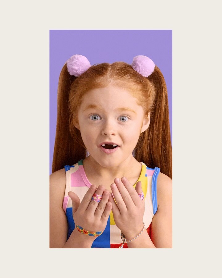 A young red-haired child with a surprised expression is centered against a violet background, showing off colorful jewelry.