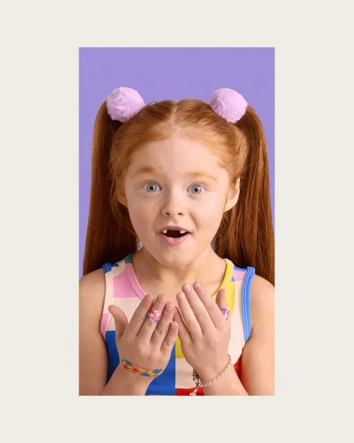 A young red-haired child with a surprised expression is centered against a violet background, showing off colorful jewelry.