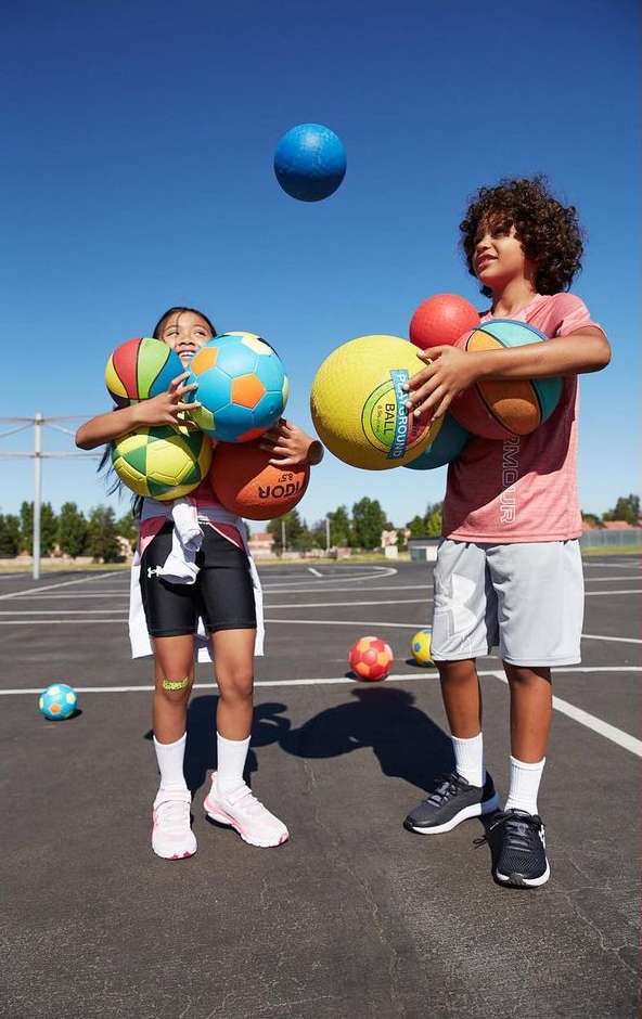 Two children, a boy and a girl, stand outdoors surrounded by colorful balls, with a clear blue sky overhead and sparse vegetation.