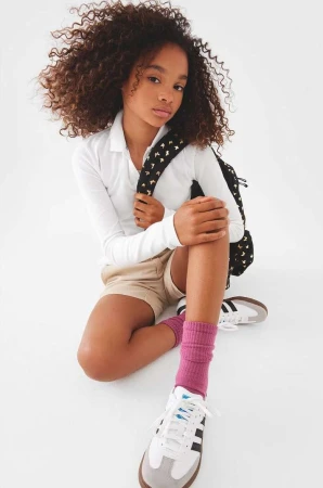 Young girl with curly hair sits with backpack, wearing a white collared shirt, tan shorts, pink socks, and white sneakers in a studio shot.