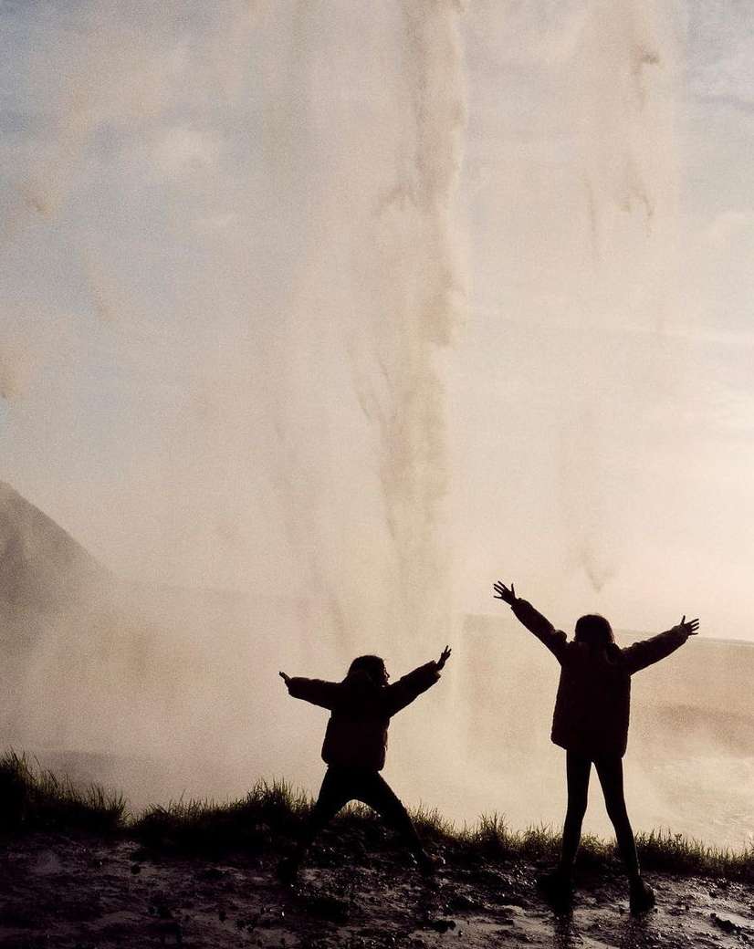 Two figures with arms raised stand before a large waterfall on a cloudy day in an outdoor setting with a neutral color palette.
