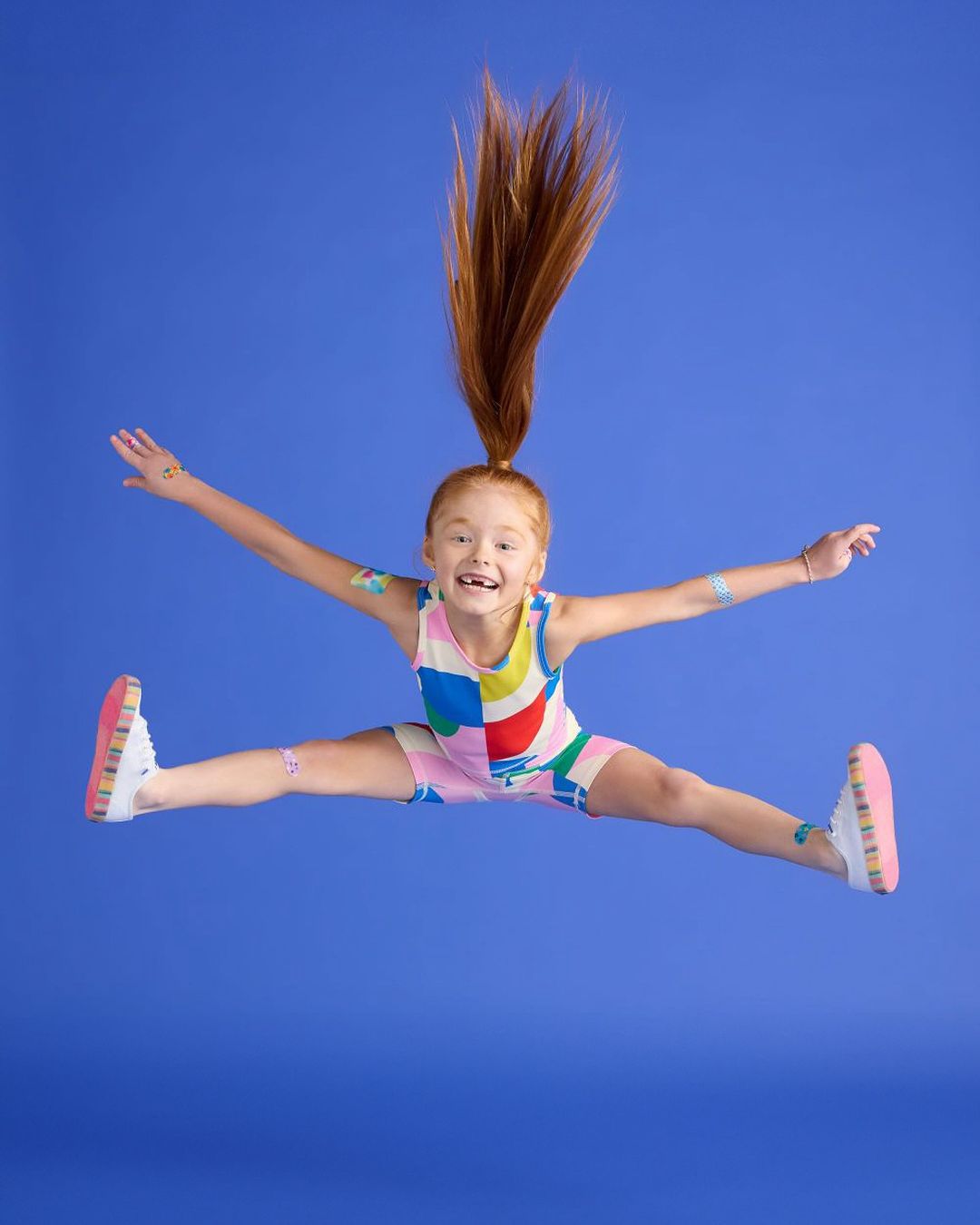 A young red-haired child jumping joyfully in mid-air against a solid blue studio backdrop wearing a colourful patterned onesie.