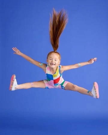 A young red-haired child jumping joyfully in mid-air against a solid blue studio backdrop wearing a colourful patterned onesie.