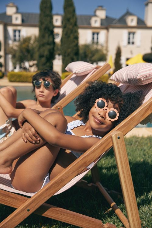 Two children, a boy and a girl, lounge in striped lounge chairs near a pool outside a large building on a sunny day.