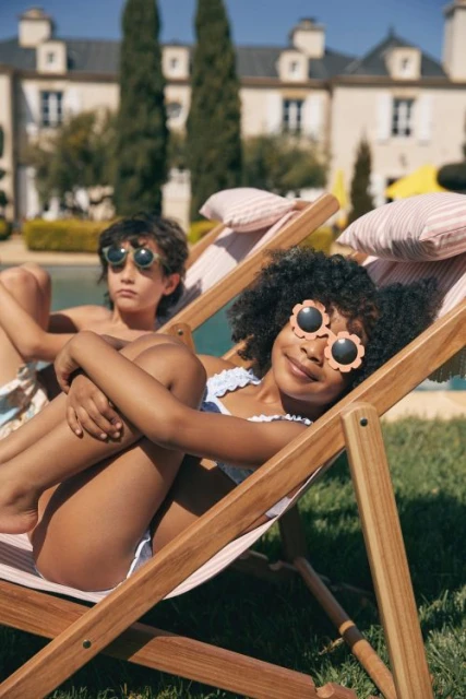 Two children, a boy and a girl, lounge in striped lounge chairs near a pool outside a large building on a sunny day.