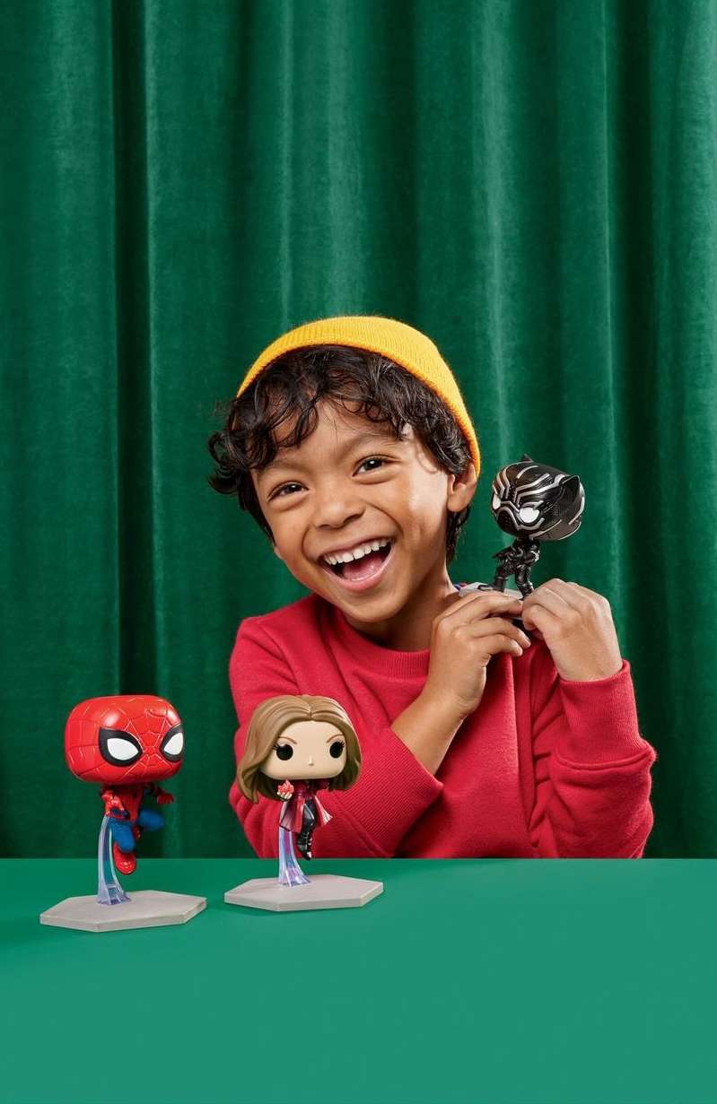 Smiling boy holds a Black Panther toy with Spiderman and Captain Marvel toys on a green table against a green curtain.