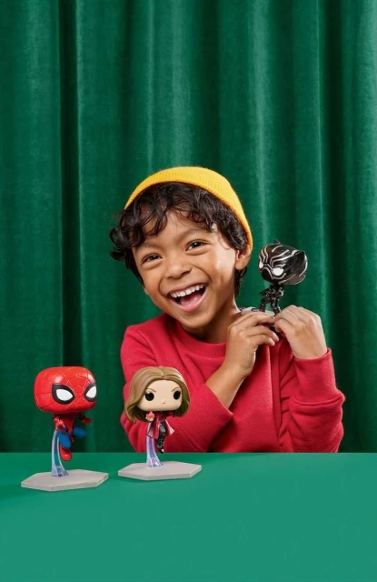 Smiling boy holds a Black Panther toy with Spiderman and Captain Marvel toys on a green table against a green curtain.