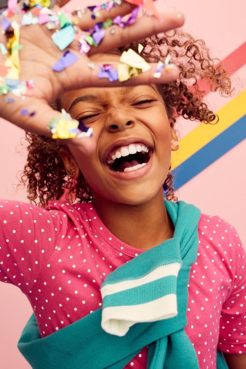 A joyful young girl with curly hair smiles and throws confetti into the air against a colorful striped background in a close-up portrait.