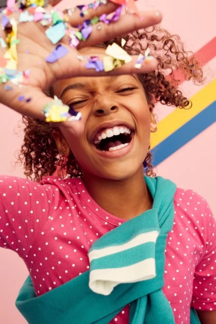 A joyful young girl with curly hair smiles and throws confetti into the air against a colorful striped background in a close-up portrait.
