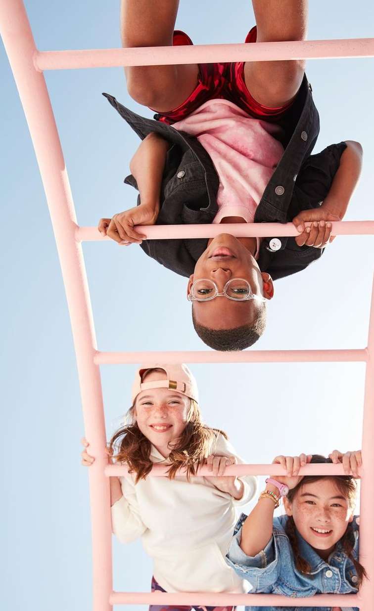 Three children play on a light-pink ladder against a bright blue sky; two girls smile on the lower rungs and a boy hangs upside down.