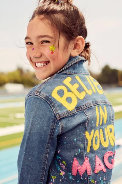 Smiling young girl with star-shaped glitter on her face poses in a denim jacket with painted words 'Believe In Your Magic'.